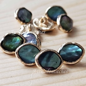 Peut inclure: Une collection de boutons ronds présentant des centres irisés bleu foncé et vert avec des bords dorés métalliques. Les boutons ont une texture de surface lisse et légèrement irrégulière ainsi qu'une attache centrale au dos. Le texte Avocadostitches est visible.