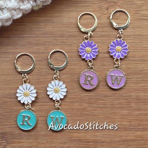 May include: Gold-colored metal earrings with daisy and circular charms. The daisy charms are white with yellow centers. The circular charms are teal and lavender, each with a letter. The text "AvocadoStitches" is at the bottom.
