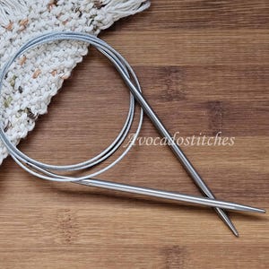 May include: Two silver knitting needles with a cable, lying on a wooden surface. The needles are crossed over each other.