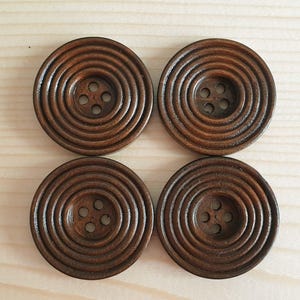 May include: Four circular dark brown wooden buttons arranged in a square formation. Each button features a series of concentric circular grooves radiating from the centre, which contains four small holes for threading, showcasing a smooth finish.