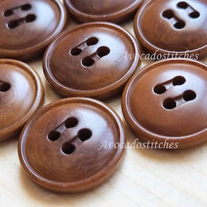 May include: A close-up of six brown wooden buttons, each with two holes. The buttons are arranged in a semi-circle on a light brown surface. The text "AvocadoStitches" is visible in the image.