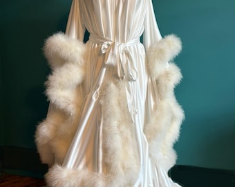 May include: A white satin dressing gown with long sleeves and a tie waist. The gown is trimmed with fluffy white feathers around the cuffs and hem. The gown is displayed on a mannequin.