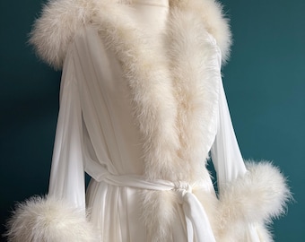 May include: A white dressing gown with feather trim on the collar and cuffs. The gown has long sleeves and a tie waist. The garment is displayed on a mannequin against a teal background.