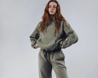 May include: A person wearing a green hooded sweatshirt and matching sweatpants. The person is standing with their hands on their hips and looking at the camera. They are wearing white sneakers.