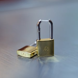 Puede incluir: Dos pendientes de candado dorados con detalles plateados. Los candados tienen las letras "AGS" grabadas.