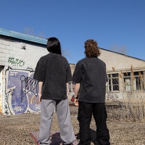May include: Two people wearing black t-shirts and pants stand on a patterned rug in front of a building with graffiti. The building has a blue trim and windows. The sky is blue.
