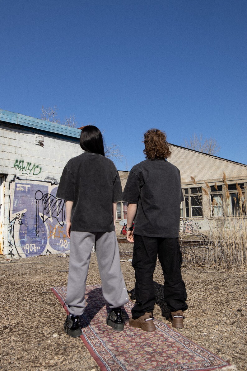 May include: Two people wearing black t-shirts and pants stand on a patterned rug in front of a building with graffiti. The building has a blue trim and windows. The sky is blue.