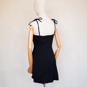 May include: A solid black mini dress featuring a straight neckline and thin shoulder straps that tie into bows at the top. The garment has a fitted bodice and a slightly flared skirt, with a visible vertical zip closure centred on the back.