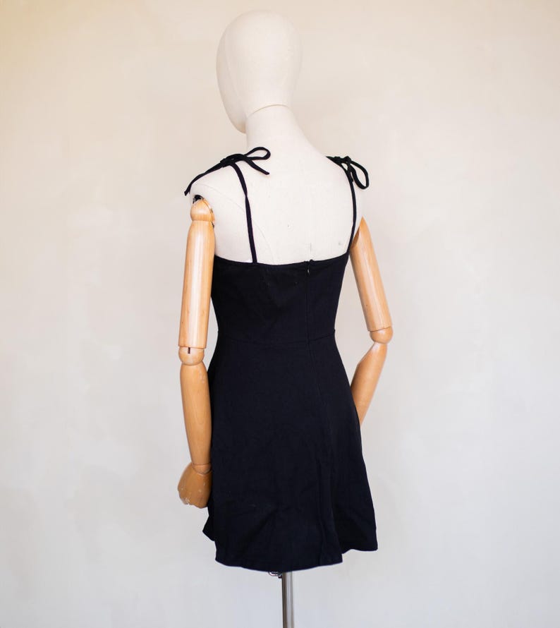 May include: A solid black mini dress featuring a straight neckline and thin shoulder straps that tie into bows at the top. The garment has a fitted bodice and a slightly flared skirt, with a visible vertical zip closure centred on the back.