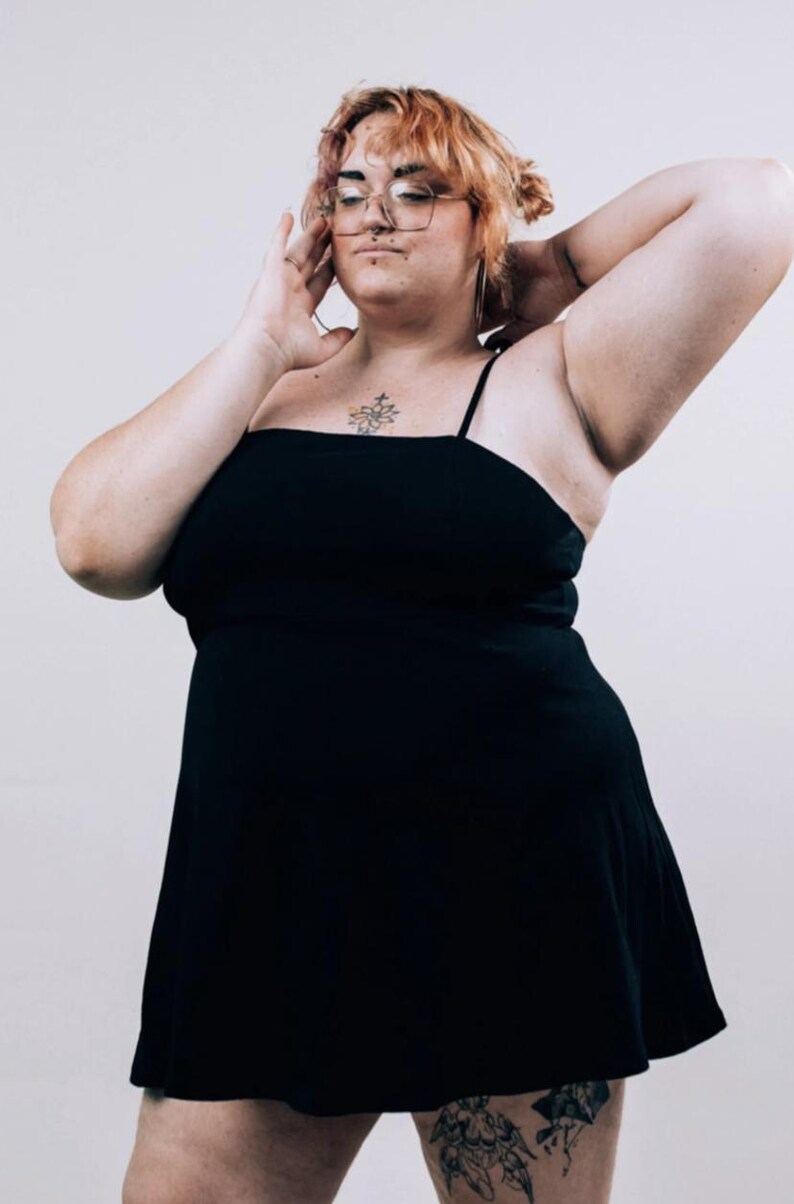 May include: A solid black mini dress featuring a straight neckline and thin spaghetti straps. The garment has a fitted bodice that transitions into a slightly flared skirt, providing a simple and clean silhouette without any additional patterns or textures.