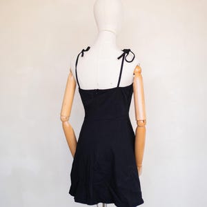 May include: A solid black mini dress featuring thin shoulder straps that tie into small bows at the top. The dress has a fitted bodice with a straight neckline and a slightly flared skirt, displayed on a mannequin with articulated wooden arms.