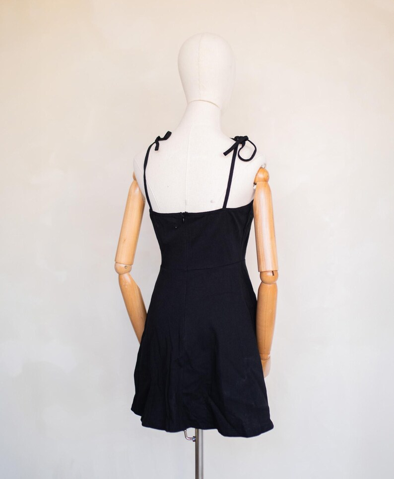 May include: A solid black mini dress featuring thin shoulder straps that tie into small bows at the top. The dress has a fitted bodice with a straight neckline and a slightly flared skirt, displayed on a mannequin with articulated wooden arms.