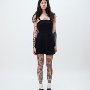 May include: A solid black mini dress featuring a straight neckline, thin spaghetti straps, and a fitted bodice that transitions into a slightly flared skirt. The garment is a simple, sleeveless piece that ends above the knee, providing a clean silhouette.