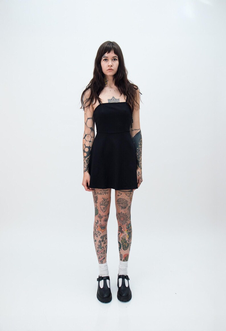 May include: A solid black mini dress featuring a straight neckline, thin spaghetti straps, and a fitted bodice that transitions into a slightly flared skirt. The garment is a simple, sleeveless piece that ends above the knee, providing a clean silhouette.