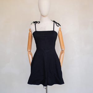 May include: A solid black mini dress featuring a square neckline and thin shoulder straps that tie into bows at the top. The garment has a fitted bodice that transitions into a flared skirt, creating a simple, structured silhouette on a mannequin.