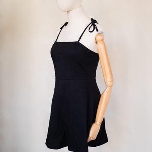 May include: A solid black mini dress featuring a square neckline and thin shoulder straps that tie into small bows at the top. The dress has a fitted bodice that transitions into a slightly flared skirt, providing a simple and clean silhouette.