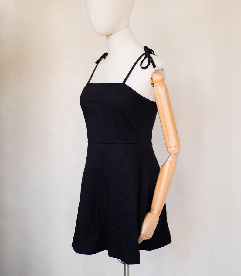 May include: A solid black mini dress featuring a square neckline and thin shoulder straps that tie into small bows at the top. The dress has a fitted bodice that transitions into a slightly flared skirt, providing a simple and clean silhouette.