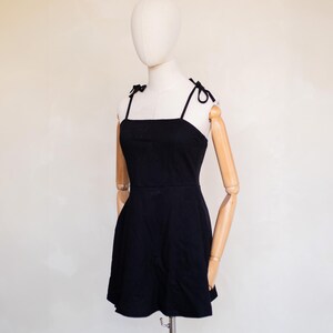 May include: A solid black mini dress featuring a square neckline and thin shoulder straps that tie into small bows at the top. The garment has a fitted bodice that transitions into a slightly flared skirt, providing a clean and simple silhouette.