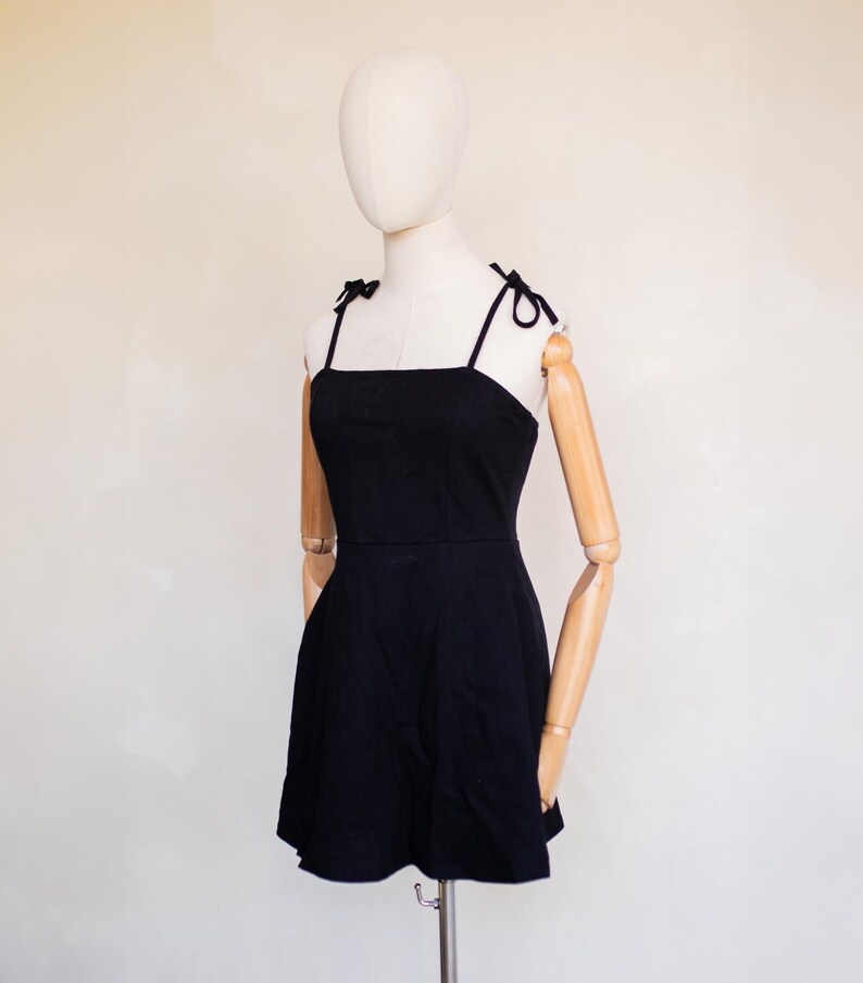 May include: A solid black mini dress featuring a square neckline and thin shoulder straps that tie into small bows at the top. The garment has a fitted bodice that transitions into a slightly flared skirt, providing a clean and simple silhouette.