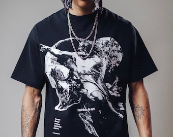 Puede incluir: Una camiseta negra con un estampado gráfico blanco de un ángel con alas y una calavera. El texto "fashion is art" está impreso en la camiseta. El modelo lleva un pantalón de estampado colorido.