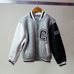 May include: A gray, white, and black varsity jacket with a large "C" on the front and an American flag on the right sleeve. The jacket has a black and white striped collar and cuffs.