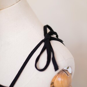 May include: A black garment strap tied into a bow on the shoulder of a cream-coloured dress form. The thin, fabric strap is knotted securely at the top of the shoulder, creating a small decorative loop detail against the neutral mannequin surface.