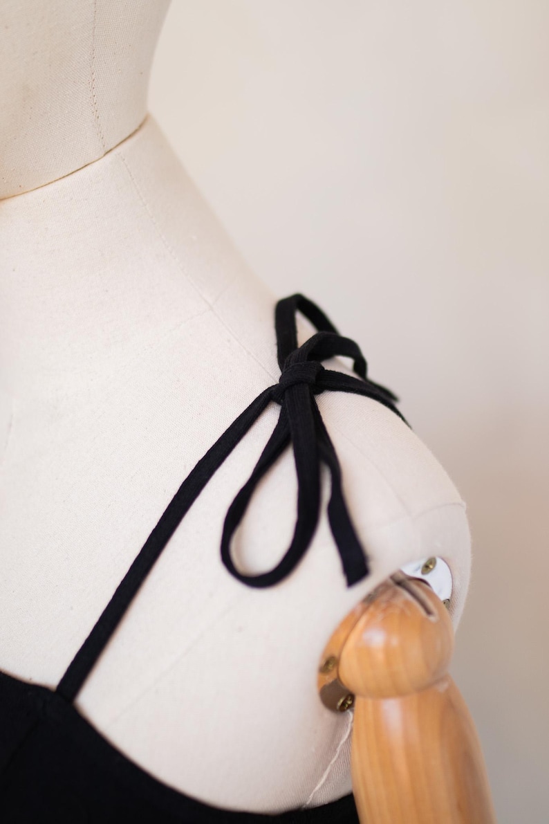 May include: A black garment strap tied into a bow on the shoulder of a cream-coloured dress form. The thin, fabric strap is knotted securely at the top of the shoulder, creating a small decorative loop detail against the neutral mannequin surface.