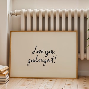 May include: A framed print with the words "Love you, goodnight!" in black script. The print has a light beige background and a natural wood frame. It is leaning against a wall near a radiator and a window.