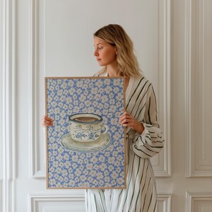 May include: A framed print of a blue and white teacup with a floral pattern on a blue background. The print is held by a person in a striped dress.