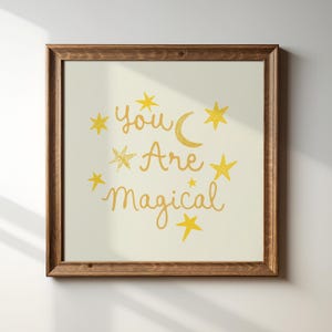 以下が含まれることがあります： 「You Are Magical」の文字が金色で書かれた、金色の星と三日月が描かれた額入りのプリント。 フレームは濃い茶色の木製で、背景はオフホワイトです。