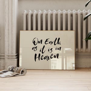 May include: A framed print with the text "On Earth as it is in Heaven" in black calligraphy. The print has a gold frame and is leaning against a white radiator. A rolled-up magazine rests on the wooden floor in front of the print.