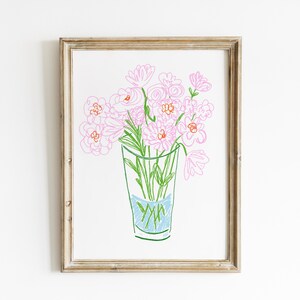 May include: A framed art print featuring a hand-drawn illustration of pink flowers in a clear glass vase. The flowers have pink petals and green stems. The vase has a blue section at the bottom. The frame is a light brown color.