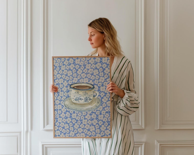 May include: A framed print of a blue and white teacup with a floral pattern on a blue background. The print is held by a person in a striped dress.