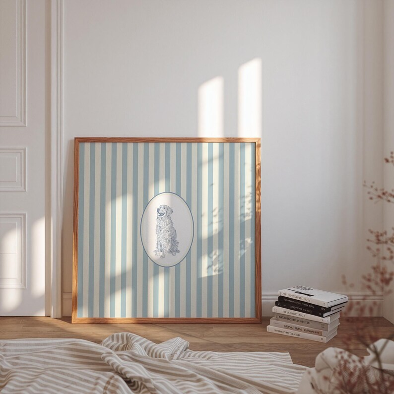 May include: A framed print featuring a dog illustration within an oval, set against a backdrop of vertical blue and cream stripes. The wooden frame complements the artwork, creating a classic aesthetic. A stack of books rests nearby.