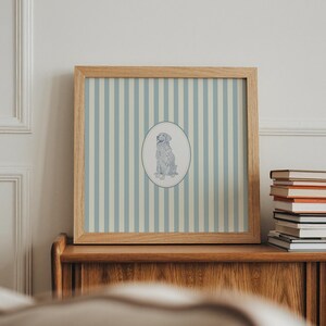 May include: A framed print featuring a dog illustration within an oval, set against a backdrop of blue and white vertical stripes. The wooden frame complements the artwork, creating a classic aesthetic. The print is displayed on a wooden cabinet, with a stack of books nearby.