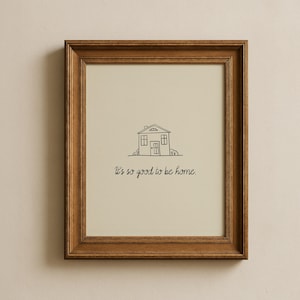 May include: A framed print with a simple line drawing of a house, complete with windows and a door. The text "It's so good to be home" is written below the house in a handwritten style. The frame is a warm brown colour.