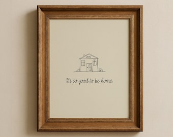 May include: A framed print featuring a minimalist line drawing of a house with a gable roof, windows, and a door. The text "It's so good to be home" is written below the house in a handwritten style. The frame is a warm brown color.