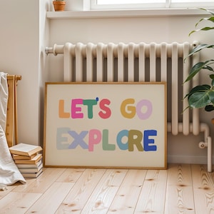Puede incluir: Un cuadro enmarcado con la frase "LET'S GO EXPLORE" en letras coloridas. La obra de arte se muestra sobre un fondo beige claro, enmarcada en madera natural, contra una pared de tono neutro y un suelo de madera.