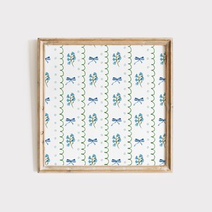 May include: A framed print with a repeating pattern of blue flowers and bows on a white background with green scalloped edges.
