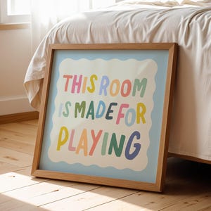 May include: A framed print with a light blue background and a white border. The text "THIS ROOM IS MADE FOR PLAYING" is written in colorful, playful letters.