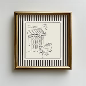 May include: Black and white illustration of a cafe with a striped awning that says "Cafe et Boulangerie". The cafe has a table with two chairs outside. The illustration is framed in a gold frame with a black and white striped background.