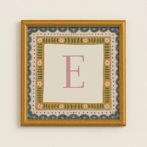 Op de afbeelding: Vierkante ingelijste kunstdruk met de letter "E" in roze op een crèmekleurige achtergrond. De gouden lijst omringt een decoratieve rand in marine, goud, roze en rood. Een charmant stukje wandkunst.