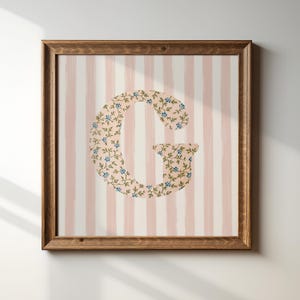 以下が含まれることがあります： ピンクと白のストライプの背景に、大きな「G」の文字が中央に配置された額装されたプリント。文字は、小さな青い花が咲いた花柄で覆われています。