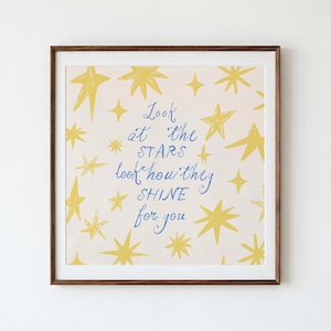 May include: A square framed wall art piece featuring a light cream background with scattered yellow hand-drawn stars. Centered in blue cursive and print text is the phrase: Look at the STARS look how they SHINE for you. The frame is dark brown wood.