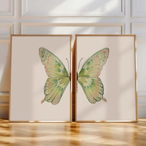 May include: Two framed butterfly art prints. Each print features a watercolor butterfly with green and pink wings, set against a beige background. The frames are gold-colored, adding a touch of elegance to the artwork. The artwork is displayed on a wooden floor.