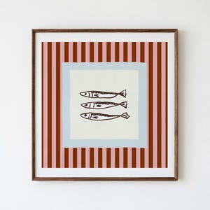 May include: A square framed wall art piece featuring a central cream-colored square with three dark brown hand-drawn fish illustrations. The central square is bordered by a light blue frame, set against a background of vertical pink and brown stripes.