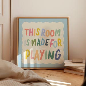 May include: A framed print with a light blue background and a white cloud-shaped border. The text "This room is made for playing" is written in colorful, playful letters.