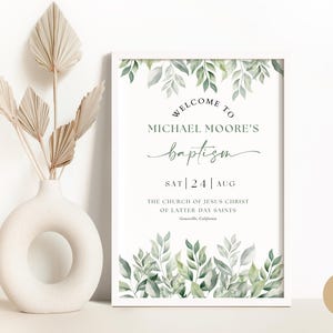 以下が含まれることがあります： 「WELCOME TO MICHAEL MOORE'S baptism SAT | 24 | AUG」のテキストが入った額入りの洗礼看板。看板は緑の葉で装飾されており、乾燥したヤシの葉が入った白い花瓶の隣にあります。