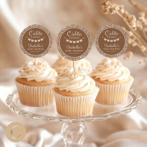 Peut inclure: Des cupcakes avec glaçage blanc et des décorations sur un présentoir à gâteau en verre. Les décorations sont marron avec le texte "Cielito Lindo" et "Isabella's Baby Shower 11.06.20XX". Les cupcakes sont dans des caissettes blanches.