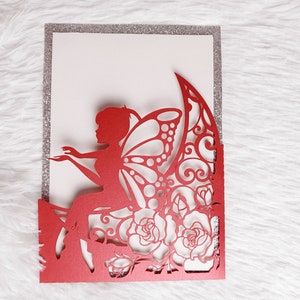 May include: Red laser cut invitation card with a fairy sitting on a crescent moon with roses and intricate designs. The card has a silver glitter border.